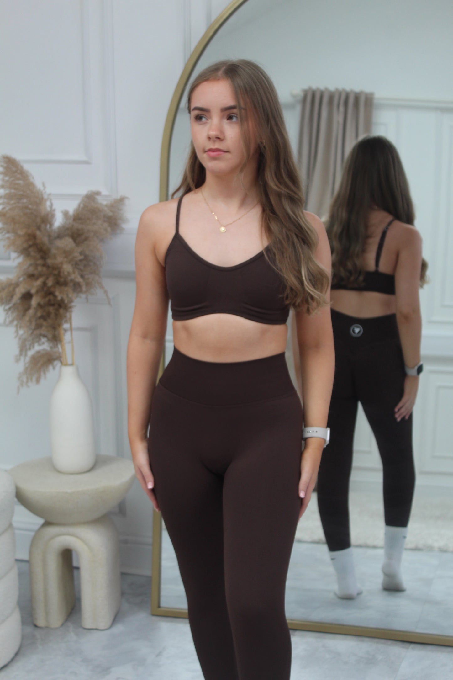 Young woman models The Eclipse Collection - Gym Bra, showcasing its halter design and flattering fit in front of an arched mirror in a minimalist room.