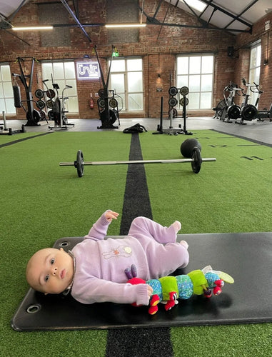 A baby lying on a black surface in a gym next to a green and blue caterpillar stuffed animal.