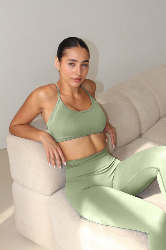 Young woman in light-green Ilume Collection sports bra, seated on a sofa, showcasing the bra's adjustable cross-back design and supportive fit.