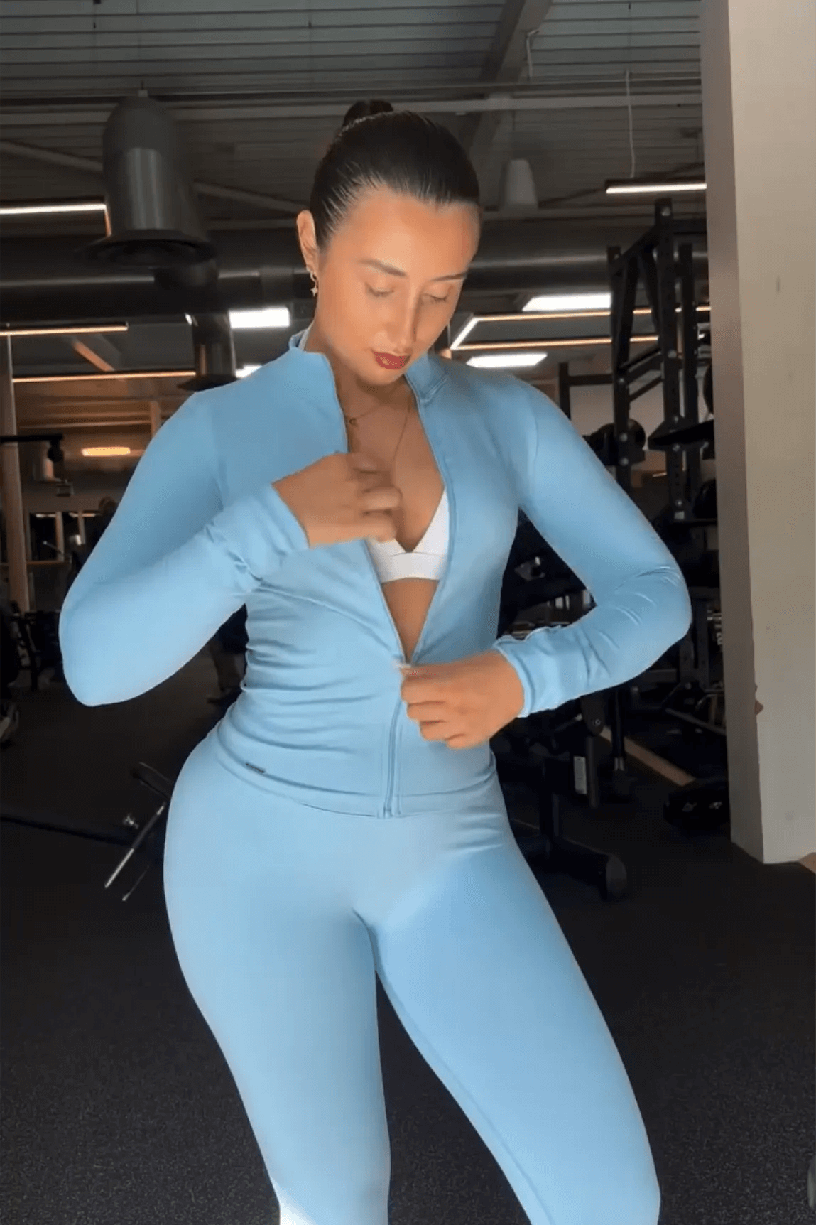 Person in gym wearing 3 Piece Set Edge Jacket, Leggings, and Ilume Top in Blue, adjusting jacket, surrounded by gym equipment, prepared for workout.