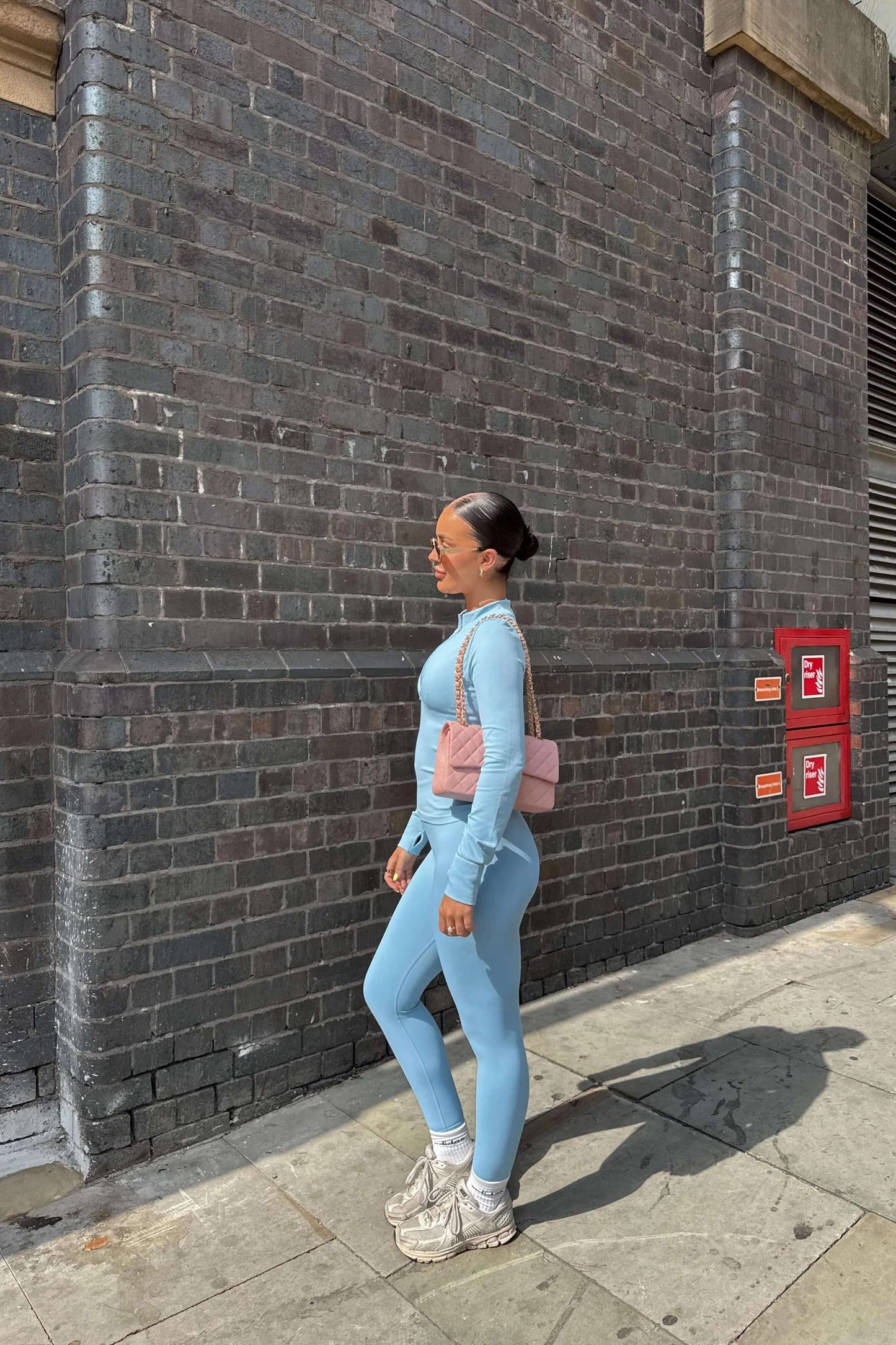Woman in profile wearing 3 Piece Set Edge Jacket, Leggings, and Ilume Top in Blue, paired with chunky sneakers and sunglasses, against a brick wall.