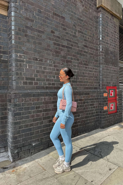 Woman in profile wearing 3 Piece Set Edge Jacket, Leggings, and Ilume Top in Blue, paired with chunky sneakers and sunglasses, against a brick wall.