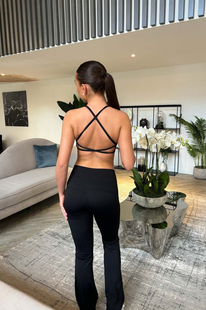 Woman wearing Pilates Reformer Collection in Black - Twist Front & Cross Back Bra, standing in a room with a couch, plants, and wall art.