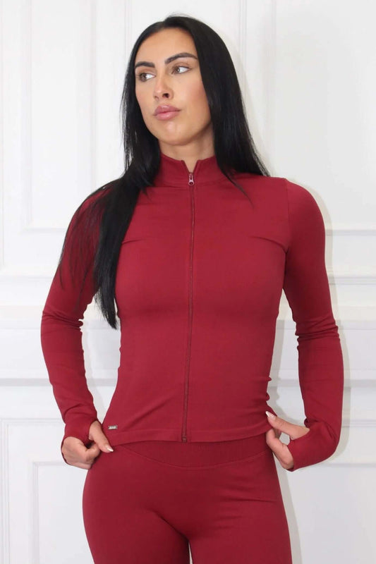 Woman wearing The Empower Edge Collection - Gym Jacket, a form-fitting, long-sleeve zip-up top, with matching high-waisted leggings, stands against a light wall.