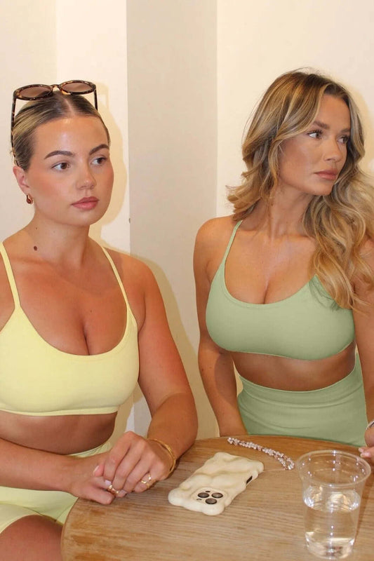 Two women in sports bras from The Ilume Collection sit at a table, highlighting the adjustable cross back design for comfort and support.