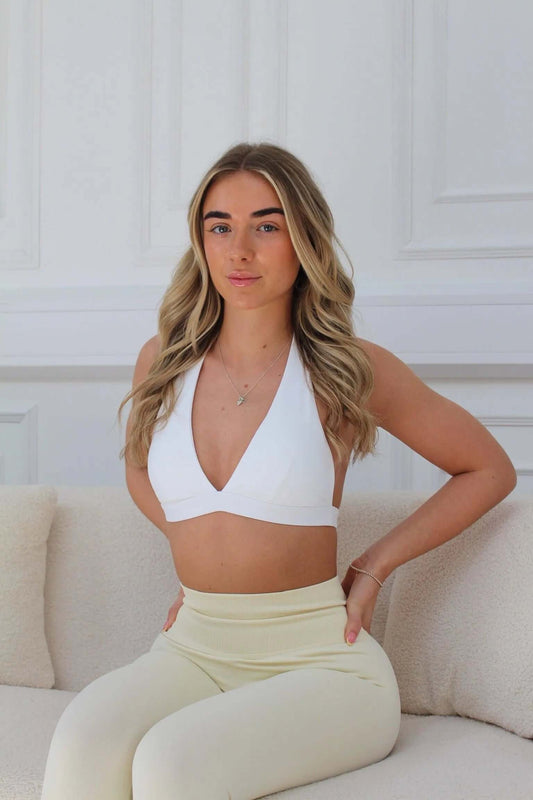 Young woman in The Ilume Collection - Halter Low Cut Gym Bra, seated on a textured sofa, showcasing the bra's flattering low-cut halter neckline and supportive design.