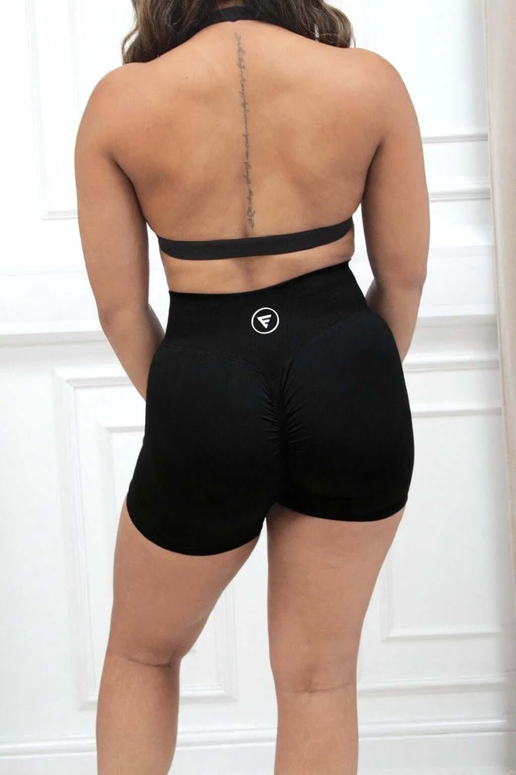 Woman in black sports bra wearing The Ilume Collection Scrunch Bum Gym Shorts, highlighting the scrunch bum design and logo at the waistband against a white wall.
