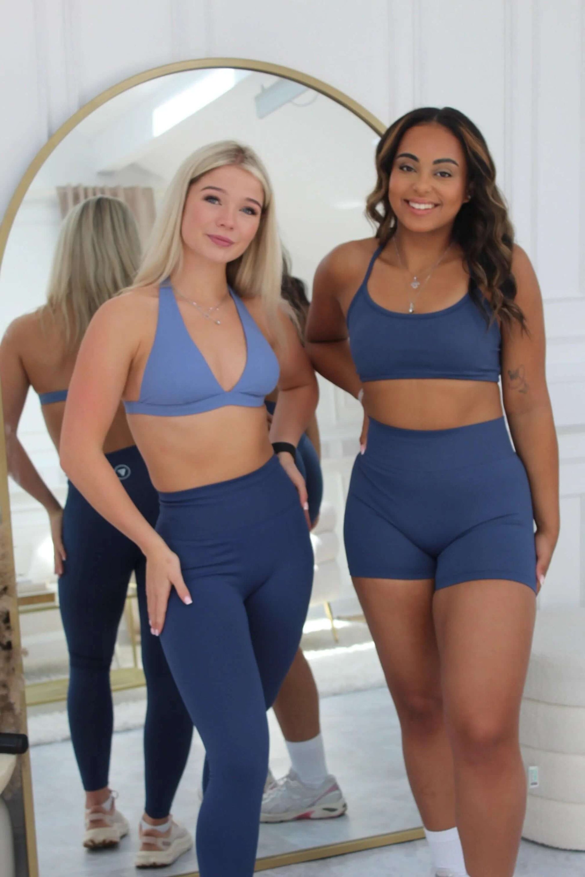 Two women in The Ilume Collection Scrunch Bum Gym Shorts, showcasing high-waisted, supportive design, smiling in front of a mirror in a minimalist room.