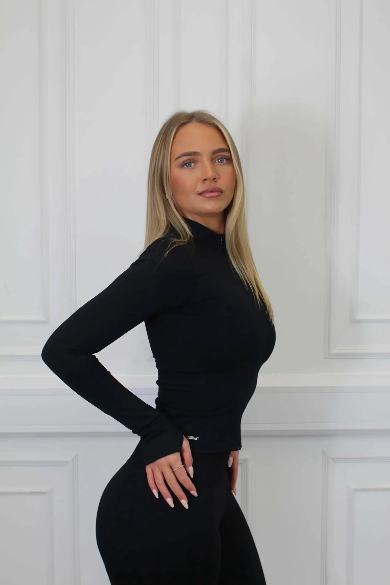 Young woman models The Empower Edge Collection - Gym Jacket, showcasing its form-fitting design, highlighting luxury, comfort, and personalized fit against a white paneled wall.